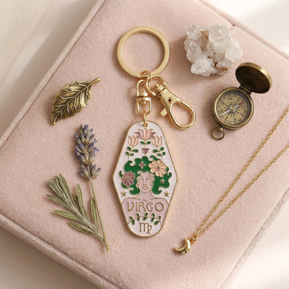 A gold-tone keychain with an enamel pendant shaped like a coffin, featuring a green and pink astrological design with the text 'Virgo MP'. The keychain is displayed on a light pink velvet jewelry box alongside decorative props including a gold leaf, lavender, a crystal cluster, a vintage-style compass, and a gold necklace.