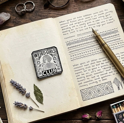 A styled flat-lay featuring a vintage-style open book with printed text, a small square pin reading 'WEIRD GIRLS CLUB', a gold pen, a cup of tea, glasses, rings, crystals, dried leaves, and tarot cards on a dark wooden surface.
