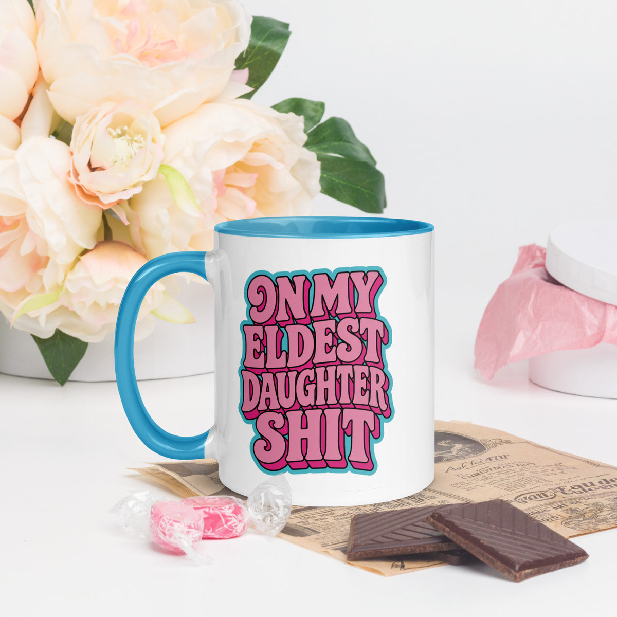 A white ceramic mug with a bright blue handle and interior, featuring the text 'ON MY ELDEST DAUGHTER SHIT' in pink bubble letters with a blue outline. The mug is styled on a tabletop with pink candy, chocolate pieces, and a vase of pale peach roses in the background.
