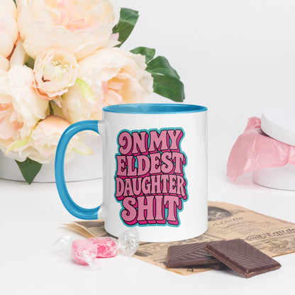 A white ceramic mug with a bright blue handle and interior, featuring the text 'ON MY ELDEST DAUGHTER SHIT' in pink bubble letters with a blue outline. The mug is styled on a tabletop with pink candy, chocolate pieces, and a vase of pale peach roses in the background.