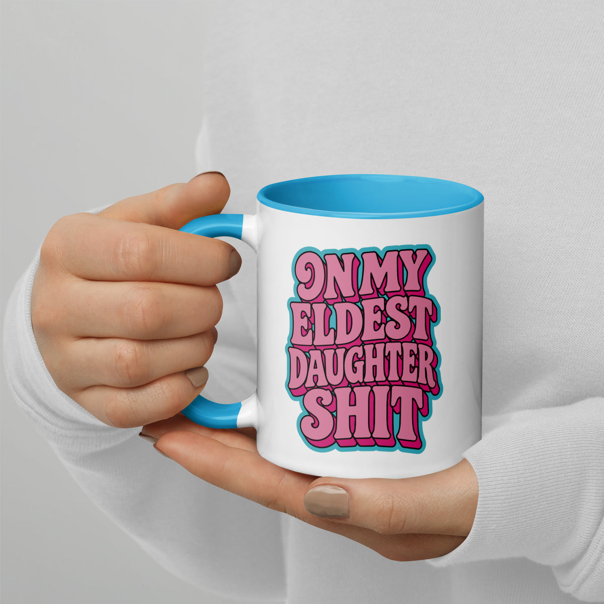 A person in a white long-sleeve shirt holds a white ceramic mug with a bright blue handle and interior. The mug features bold pink retro-style text reading 'ON MY ELDEST DAUGHTER SHIT' with a blue outline.