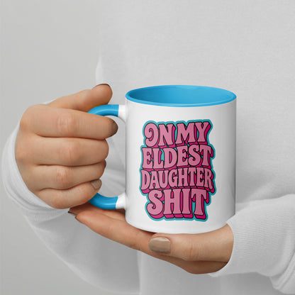 A person in a white long-sleeve shirt holds a white ceramic mug with a bright blue handle and interior. The mug features bold pink retro-style text reading 'ON MY ELDEST DAUGHTER SHIT' with a blue outline.