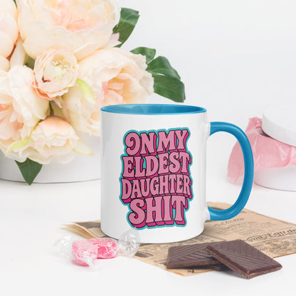 A white ceramic mug with a bright blue handle and interior, featuring the text 'ON MY ELDEST DAUGHTER SHIT' in pink bubble letters with a blue outline. The mug is styled on a white surface with decorative props, including a white vase of pale pink roses, chocolate pieces, and pink candy.