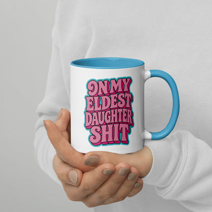 A person in a white sweatshirt holds a white ceramic mug with a blue interior and handle. The mug features a bold graphic in pink bubble letters outlined in black that reads, 'ON MY ELDEST DAUGHTER SHIT'.