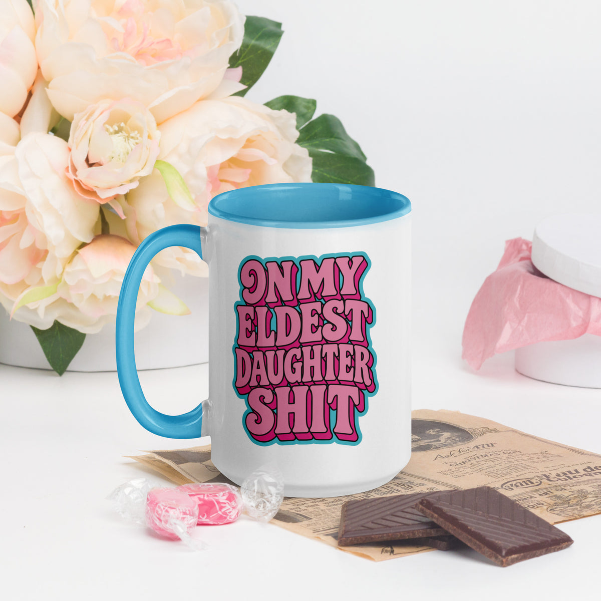 A white ceramic mug with a bright blue handle and interior, featuring the text 'ON MY ELDEST DAUGHTER SHIT' in pink and blue bubble letters. The mug is styled on a white surface with a vintage newspaper, chocolate pieces, pink candy, and a bouquet of pale peach roses in the background.