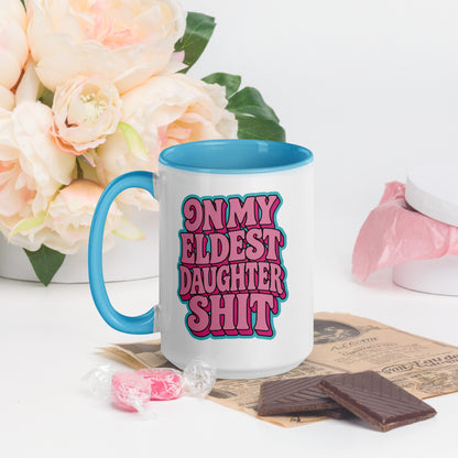 A white ceramic mug with a bright blue handle and interior, featuring the text 'ON MY ELDEST DAUGHTER SHIT' in pink and blue bubble letters. The mug is styled on a white surface with a vintage newspaper, chocolate pieces, pink candy, and a bouquet of pale peach roses in the background.