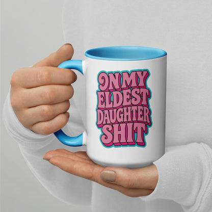 A person holding a white ceramic mug with a bright blue handle and interior. The mug features a bold graphic in pink and blue text that reads, 'ON MY ELDEST DAUGHTER SHIT'.