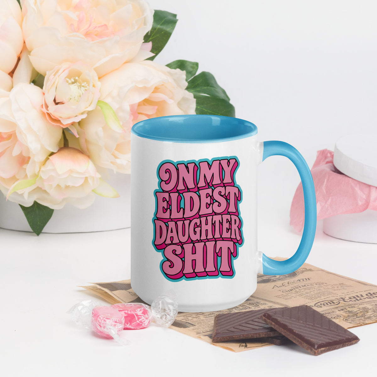 A white ceramic mug with a bright blue handle and interior, featuring the text 'ON MY ELDEST DAUGHTER SHIT' in pink and blue bubble letters. The mug is styled on a white surface with pink flowers, chocolate pieces, and candy in the background.