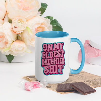 A white ceramic mug with a bright blue handle and interior, featuring the text 'ON MY ELDEST DAUGHTER SHIT' in pink and blue bubble letters. The mug is styled on a white surface with pink flowers, chocolate pieces, and candy in the background.