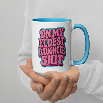 A person holding a white ceramic mug with a bright blue handle and interior. The mug features a bold graphic text that reads 'ON MY ELDEST DAUGHTER SHIT' in pink bubble letters with a teal outline.
