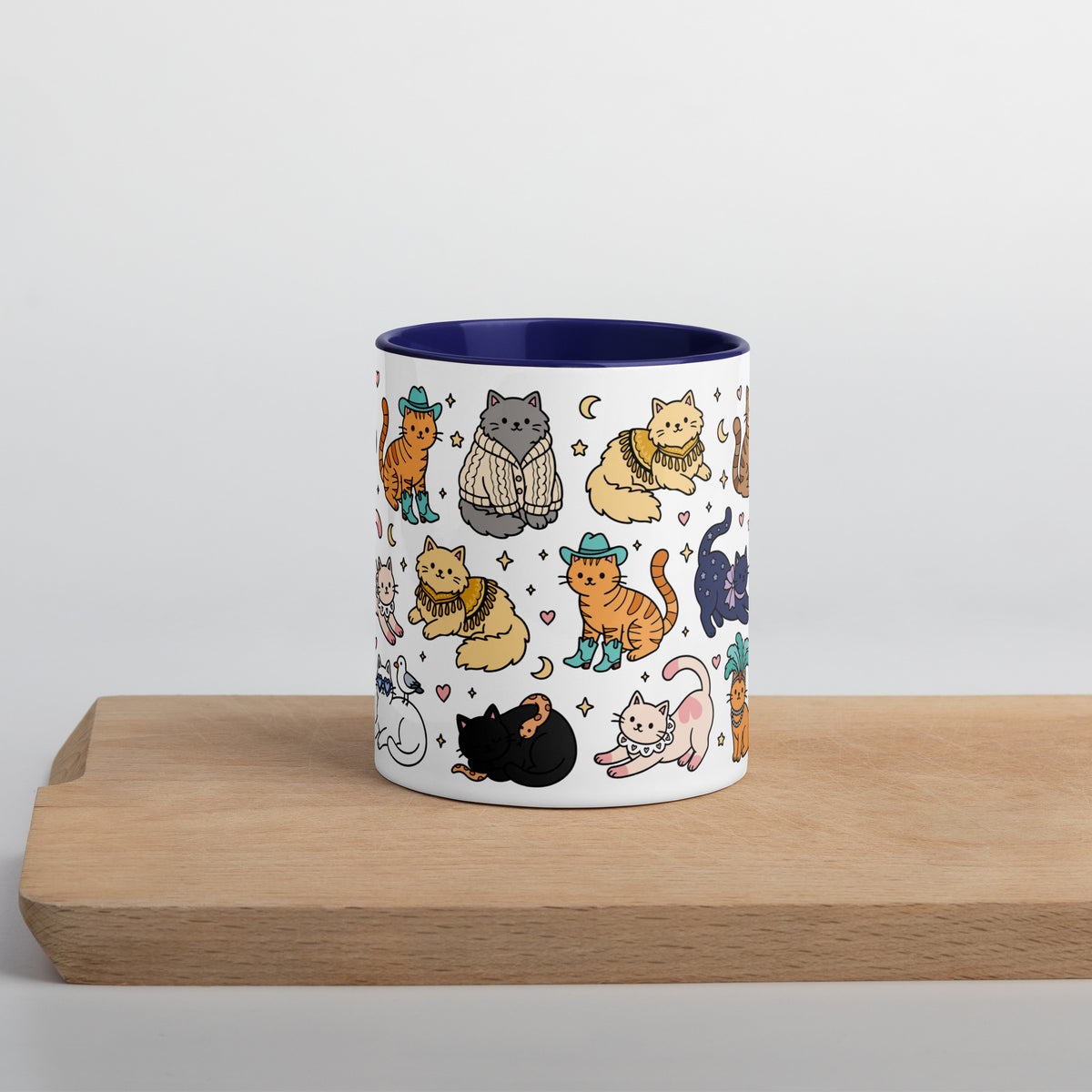 A white ceramic mug with a dark blue interior, decorated with a repeating pattern of cute hand-drawn cats in various outfits, resting on a light wooden board against a neutral light gray background.