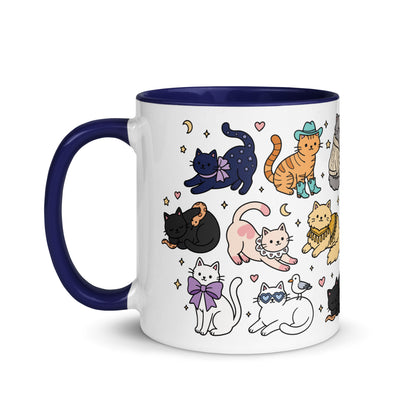 A white ceramic mug with a dark navy blue handle and interior, decorated with a whimsical pattern of various cartoon cats in different poses and outfits.