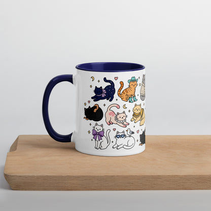 A white ceramic mug with a dark blue handle and interior, decorated with a cute pattern of various cartoon cats in different poses and outfits. The mug is shown sitting on a light-colored wooden board against a neutral light gray background.