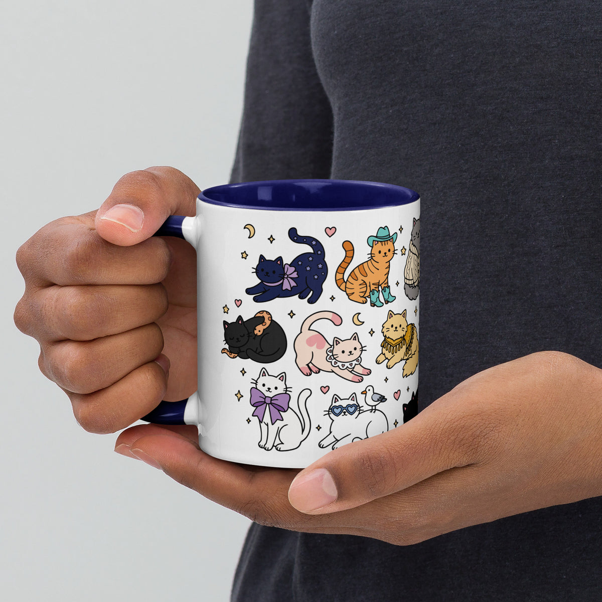 A person holding a white ceramic mug with a dark blue interior and handle, decorated with a whimsical illustration of various cats in different poses and outfits.