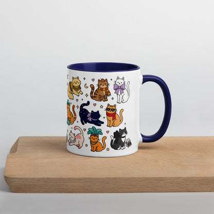 A white ceramic mug with a dark navy blue handle and interior, decorated with a colorful pattern of cartoon cats in various poses. The mug is shown sitting on a light wooden cutting board against a clean, light gray background.