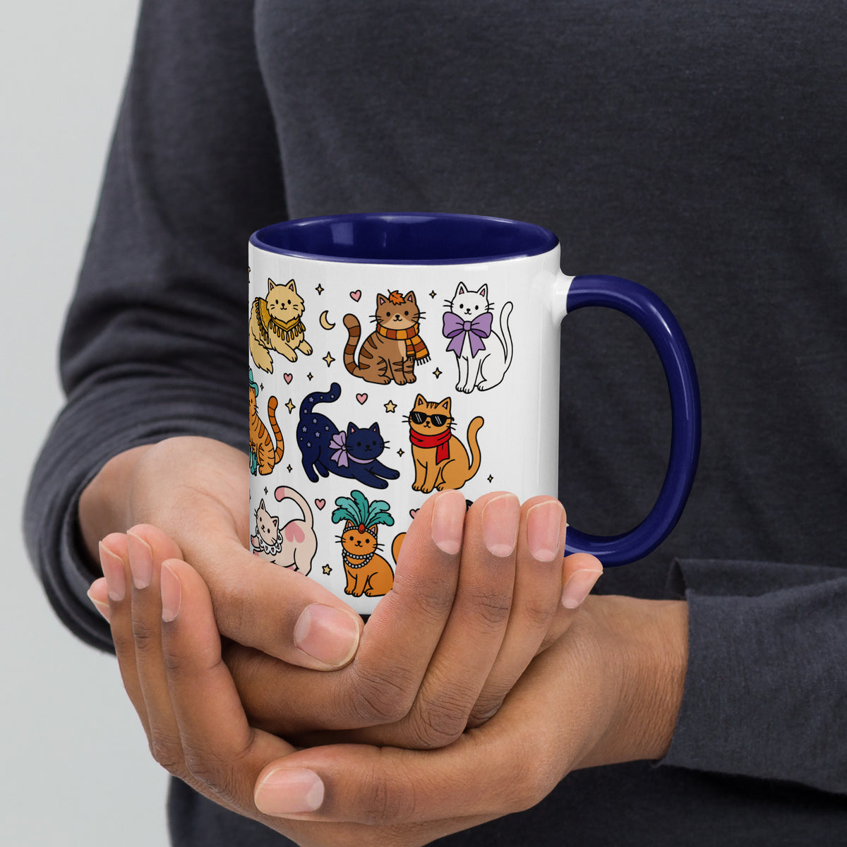 A person holding a white ceramic mug with a dark blue interior and handle, decorated with a colorful cartoon pattern of various cats.