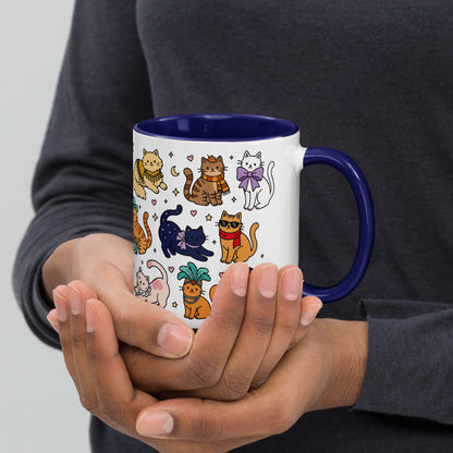 A person holding a white ceramic mug with a dark blue interior and handle, decorated with a colorful cartoon pattern of various cats.