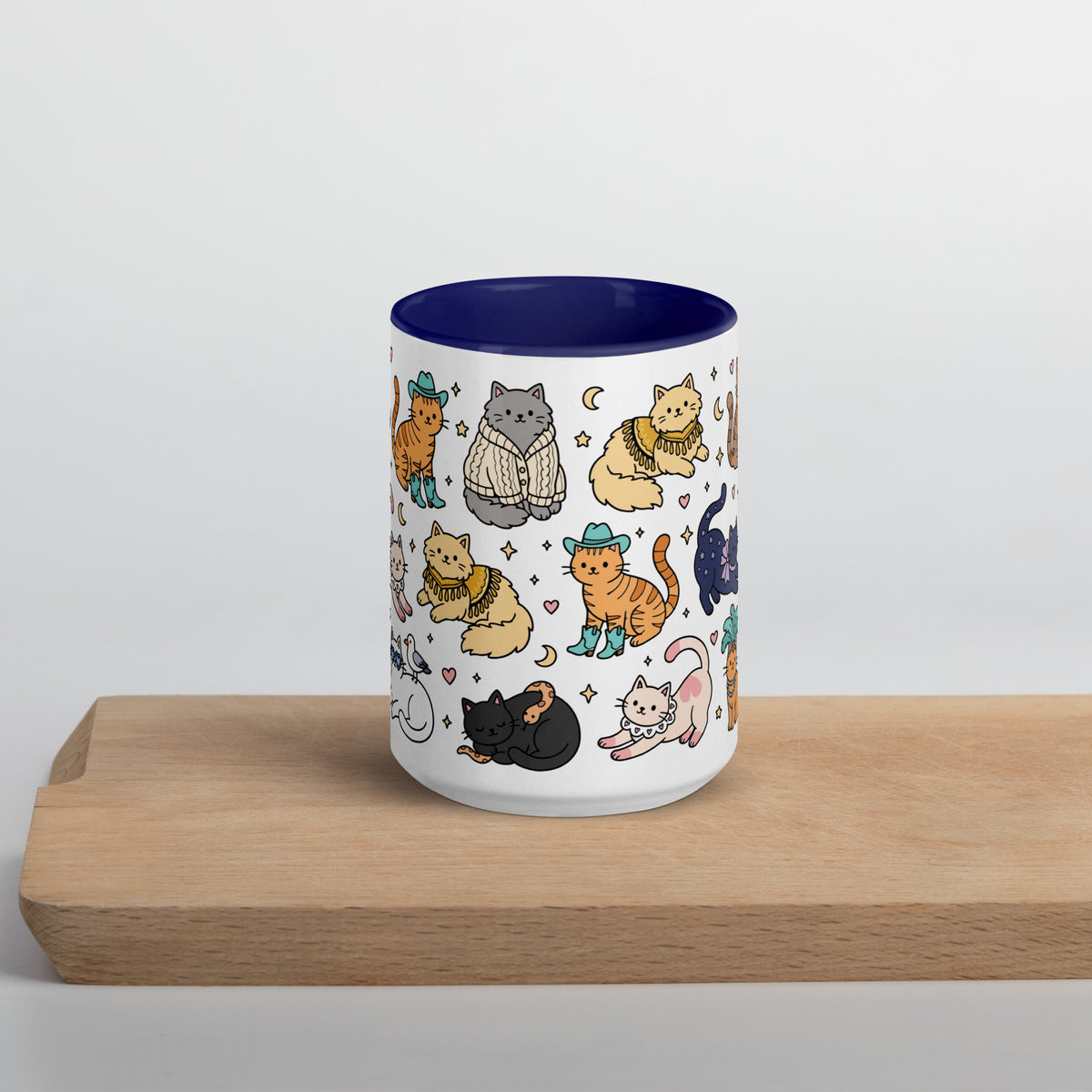 A white ceramic mug with a dark blue interior, decorated with a pattern of various cartoon cats in cozy outfits, sitting on a light wooden block against a neutral light gray background.