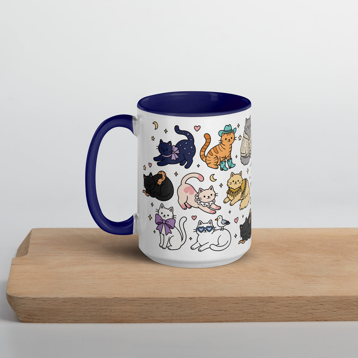 A white ceramic mug with a dark blue handle and interior, decorated with a whimsical pattern of various cartoon cats in different poses. The mug is displayed on a light wooden block against a neutral light gray background.