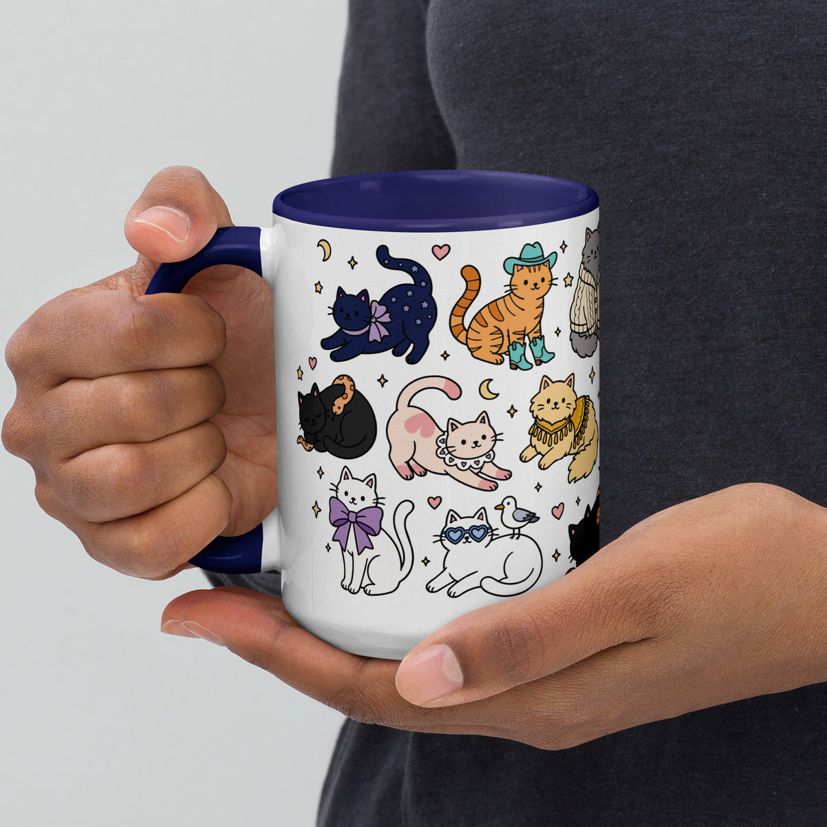 A person holds a white ceramic mug with a dark blue handle and interior. The mug is decorated with a whimsical pattern of various cartoon cats in different poses and colors, including black, orange, pink, yellow, and white, with small stars, moons, and hearts.