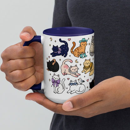 A person holds a white ceramic mug with a dark blue handle and interior. The mug is decorated with a whimsical pattern of various cartoon cats in different poses and colors, including black, orange, pink, yellow, and white, with small stars, moons, and hearts.