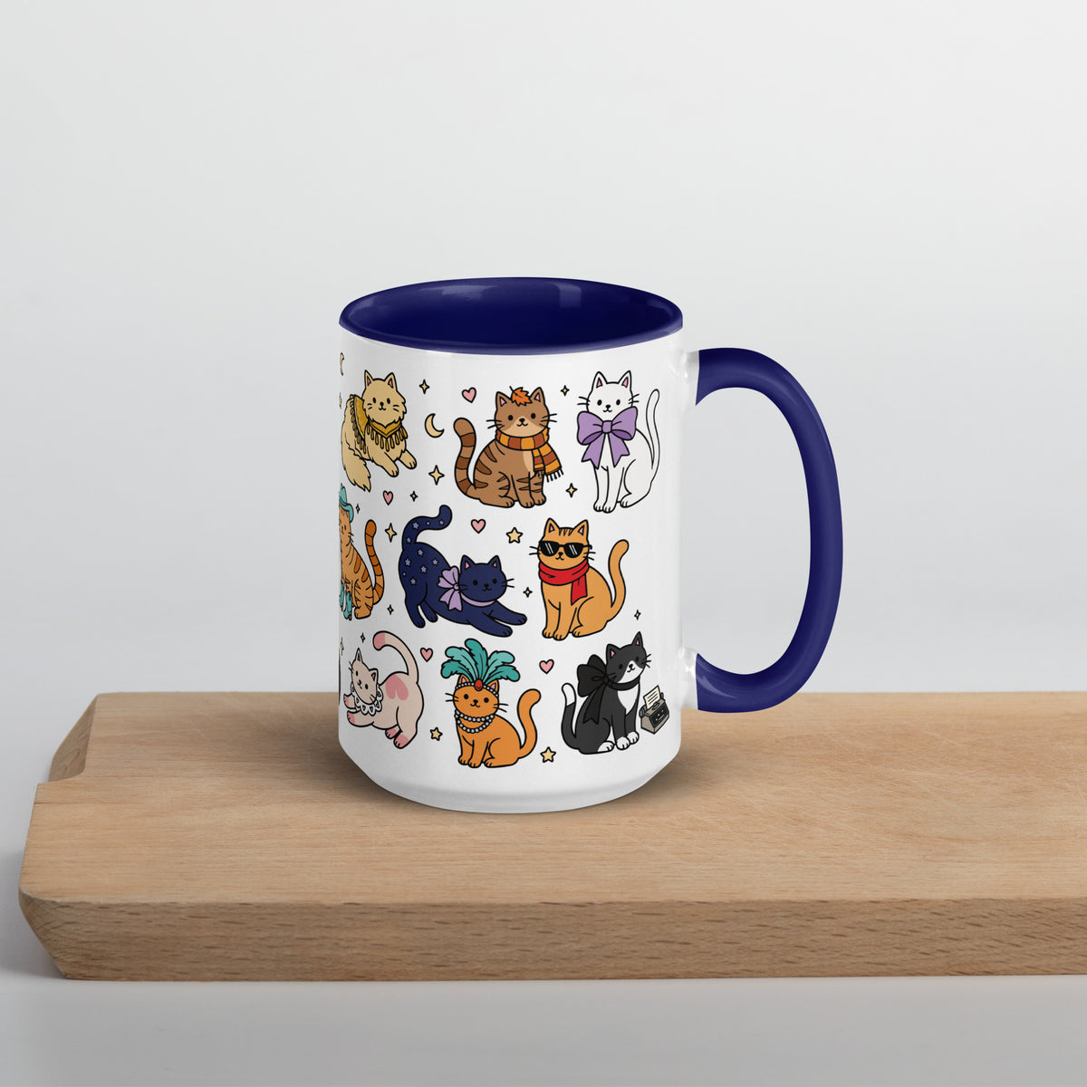 A white ceramic mug with a dark blue interior and handle, decorated with a colorful cartoon pattern of various cats, sitting on a light wooden board against a neutral light gray background.