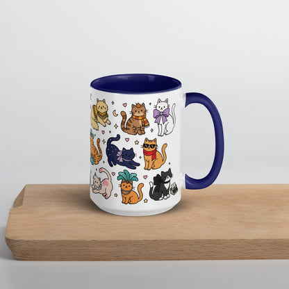 A white ceramic mug with a dark blue interior and handle, decorated with a colorful cartoon pattern of various cats, sitting on a light wooden board against a neutral light gray background.