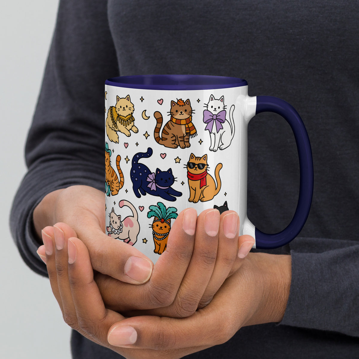 A white ceramic mug with a dark blue handle and interior, decorated with a colorful pattern of cartoon cats in various poses. The mug is being held by two hands against a person's dark gray long-sleeve shirt.