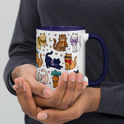 A white ceramic mug with a dark blue handle and interior, decorated with a colorful pattern of cartoon cats in various poses. The mug is being held by two hands against a person's dark gray long-sleeve shirt.