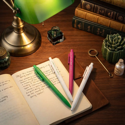 Three retractable ballpoint pens, two green and one pink, resting on an open notebook on a wooden desk next to a green lamp, books, and other desk accessories.