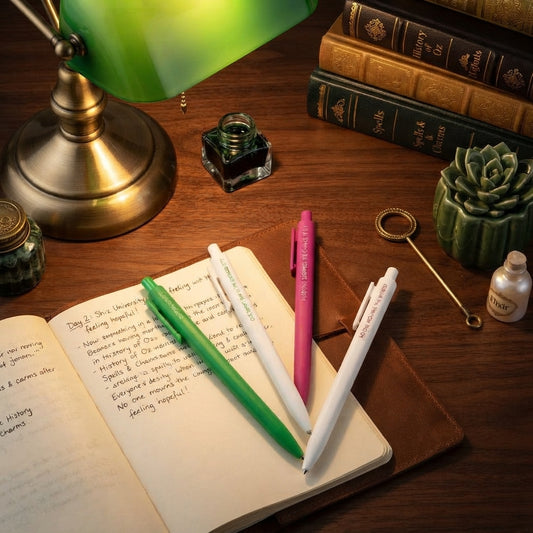 Three retractable ballpoint pens, two green and one pink, resting on an open notebook on a wooden desk next to a green lamp, books, and other desk accessories.