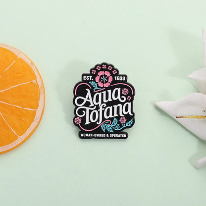 A square enamel pin with a black background and colorful floral details, featuring the text 'Aqua Tofana' and 'EST. 1833' and 'WOMAN-OWNED & OPERATED'.