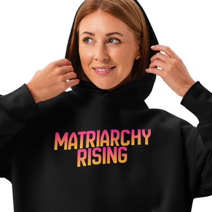 Woman wearing a black Matriarchy Rising hoodie, holding the hood, gradient lettering visible.