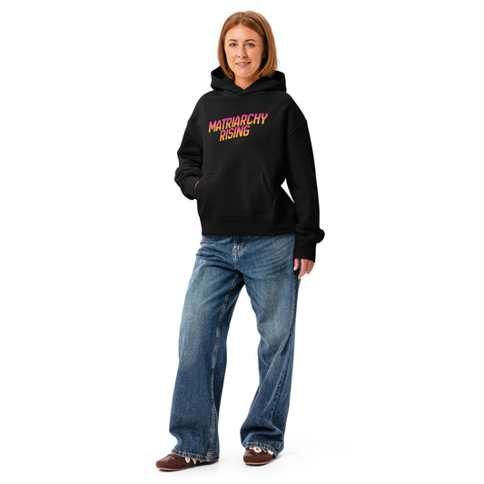 Person wearing a black Matriarchy Rising hoodie with jeans, sunrise-gradient slogan on the front.