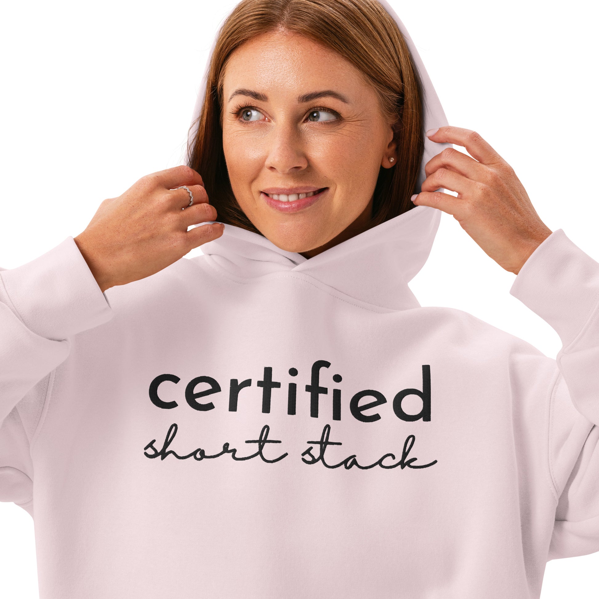 A woman wearing a light pink hoodie with the words 'certified short stack' printed in black across the chest, shown against a plain white background.